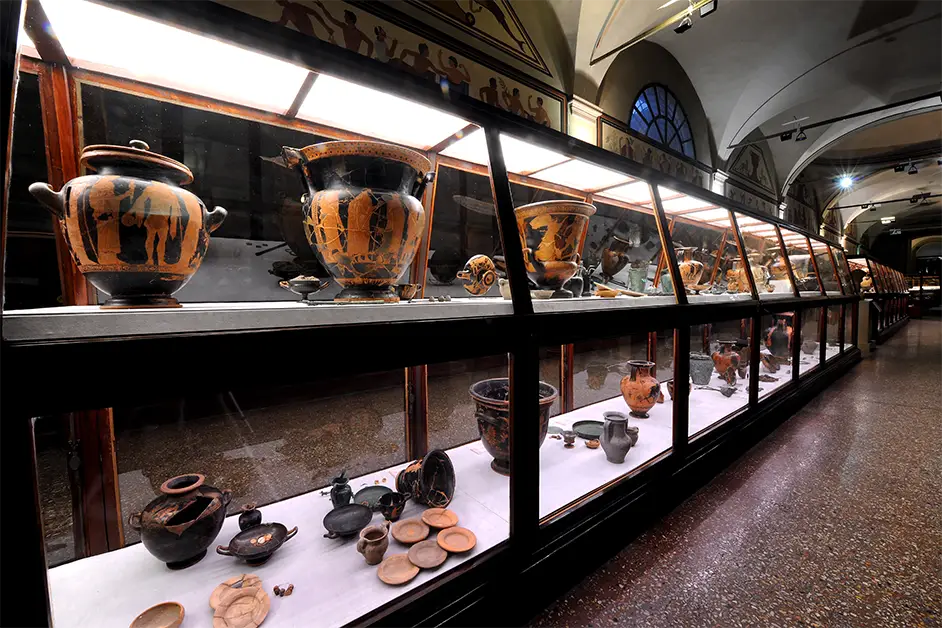 Vitrines do Museo Civico Archeologico de Bolonha exibindo cerâmicas e artefatos etruscos e gregos em uma galeria de teto abobadado.