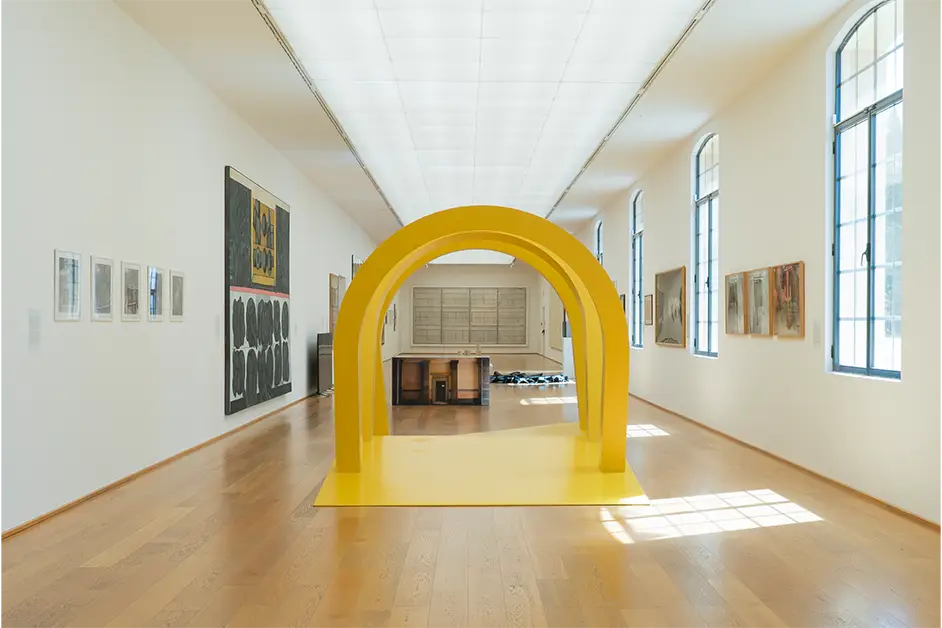 Sala iluminada do Museo d’Arte Moderna di Bologna com esculturas contemporâneas, incluindo uma estrutura amarela em arco, e obras de arte moderna nas paredes.