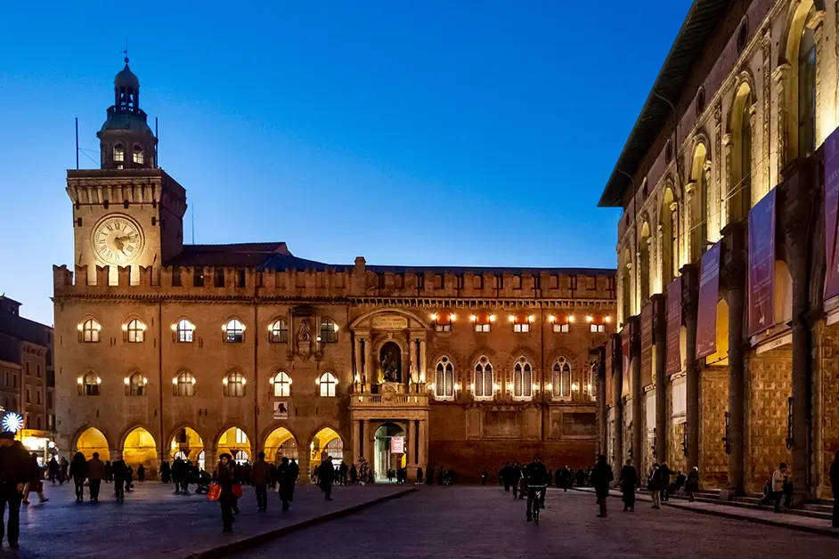 Piazza Maggiore em Bolonha ao entardecer, com o Palazzo d’Accursio iluminado e pessoas caminhando pela praça histórica.
