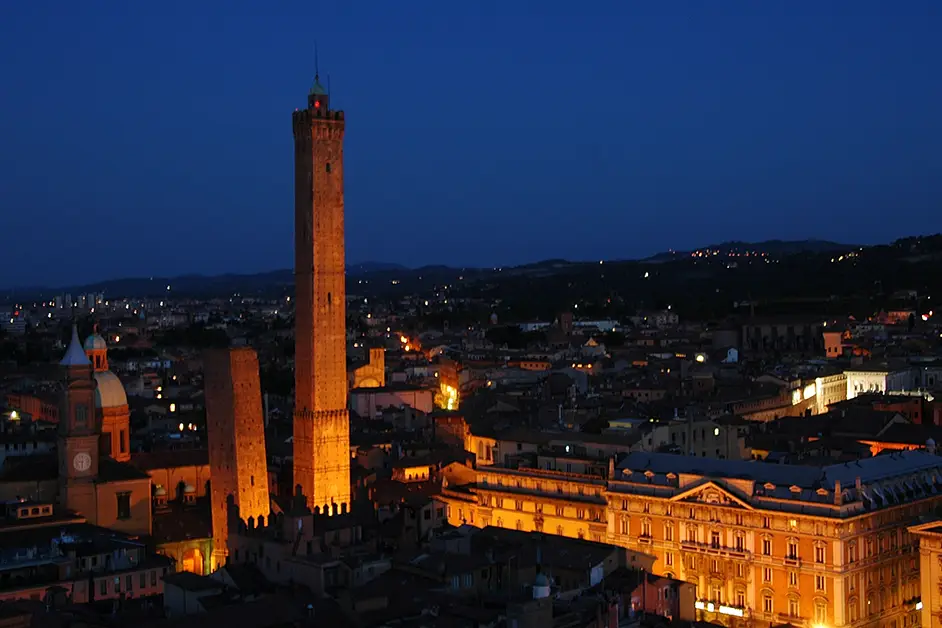 As Torres Garisenda e Asinelli iluminadas ao anoitecer, destacando-se na paisagem urbana de Bolonha com o céu azul profundo ao fundo.