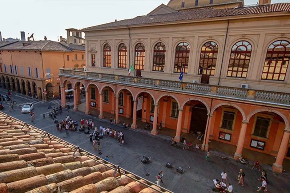 Fachada histórica da Universidade de Bolonha com colunatas em tom terracota, janelas arqueadas e movimento de estudantes na praça em frente.