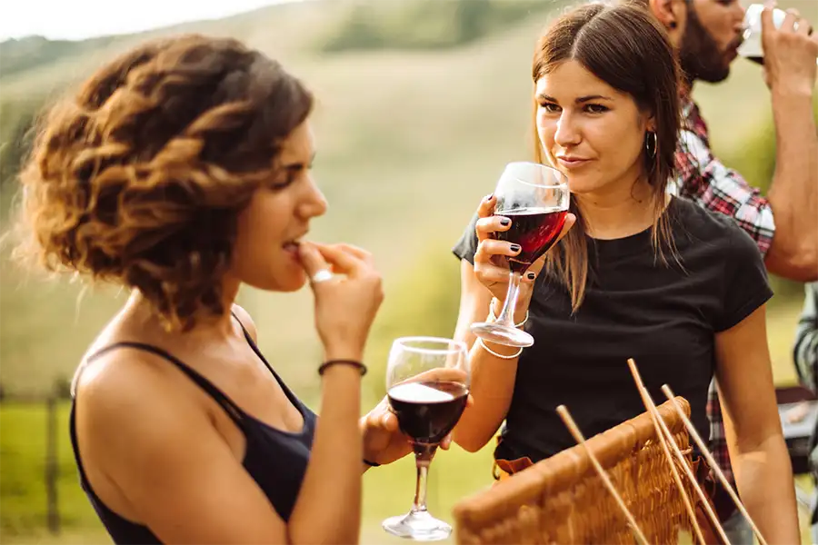 Grupo de amigos ao ar livre degustando vinho tinto; uma mulher bebe, outra prova um petisco, com paisagem de colinas ao fundo.