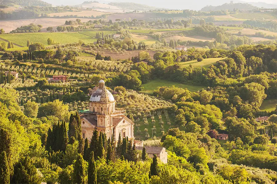 Vista panorâmica de Montepulciano, na Toscana, com igreja histórica em primeiro plano cercada por vegetação exuberante, oliveiras e colinas cultivadas ao fundo.