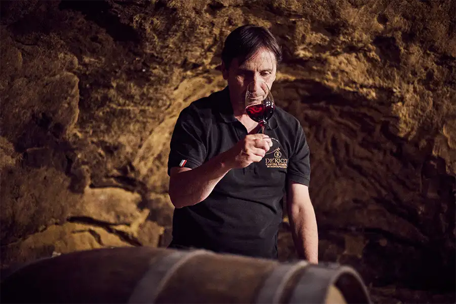 Homem em uma adega de pedra observando a coloração de um vinho tinto em uma taça, ao lado de um barril de madeira, vestindo camiseta preta com logotipo da vinícola De’ Ricci.