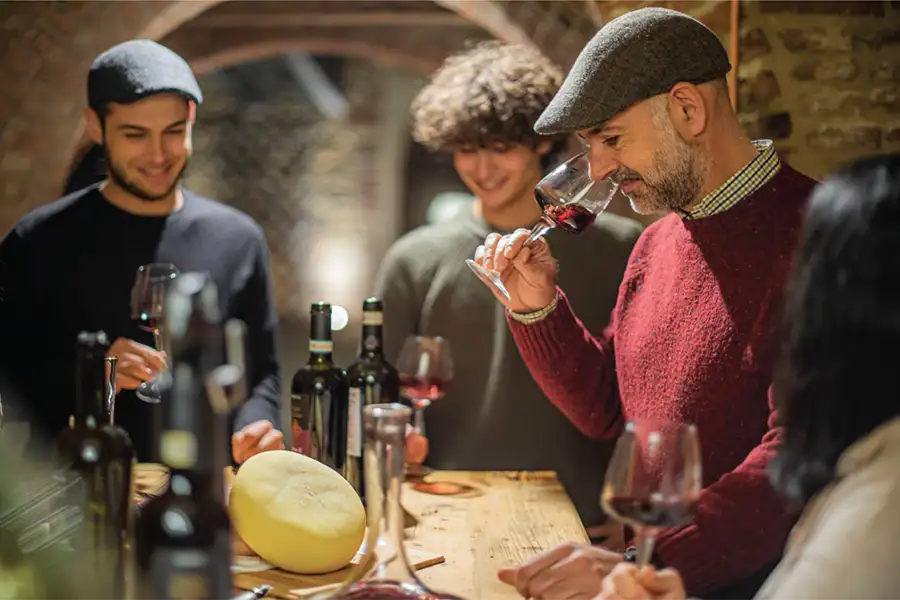 Grupo de pessoas em uma adega rústica participando de degustação de vinhos, com garrafas e queijos sobre a mesa; um homem aprecia o aroma do vinho tinto em sua taça.