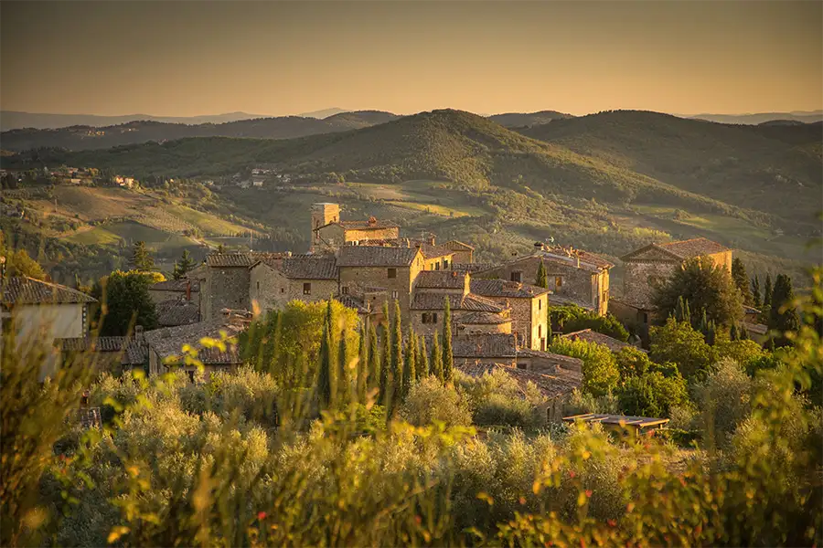 Vista ao entardecer da vila medieval de Volpaia, na Toscana, cercada por colinas verdes, ciprestes e plantações, com luz dourada iluminando as construções de pedra.