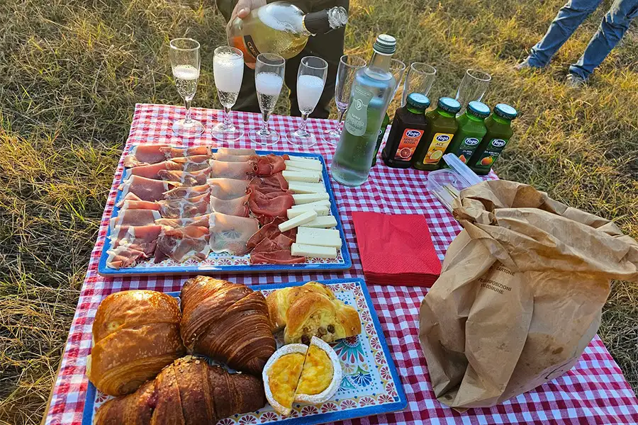 Outdoor breakfast with cold cuts, cheeses, croissants, juices, and sparkling wine on a red checkered tablecloth, after a balloon ride in Tuscany
