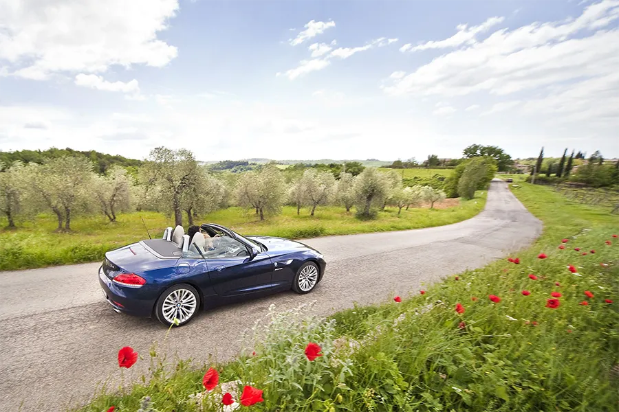 Carro conversível azul percorrendo estrada cênica entre oliveiras e flores silvestres na paisagem rural da Toscana