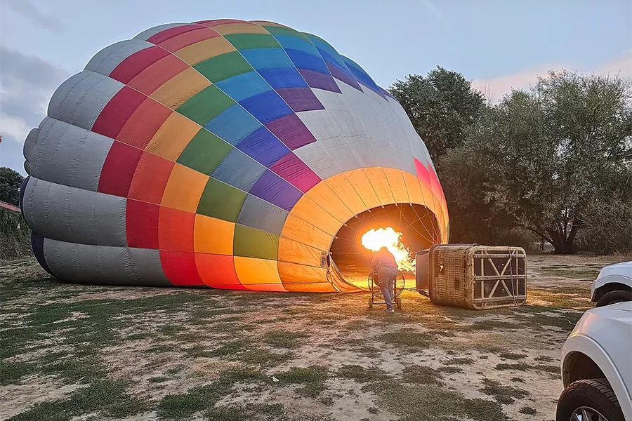 Multicolored hot air balloon being inflated with flame at dawn, with the basket positioned nearby in an open field in Tuscany