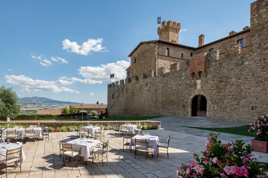 Mesas ao ar livre no pátio de pedra do restaurante La Sala dei Grappoli, em Montalcino, com vista para vinhedos e castelo medieval