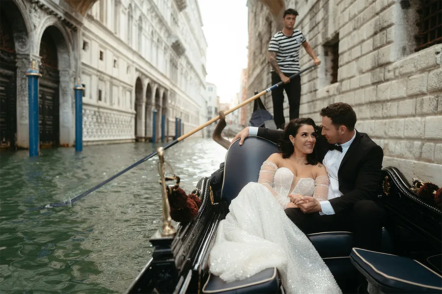 Casal elegante em passeio de gôndola por canal de Veneza, com trajes de casamento e gondoleiro ao fundo, em clima romântico.